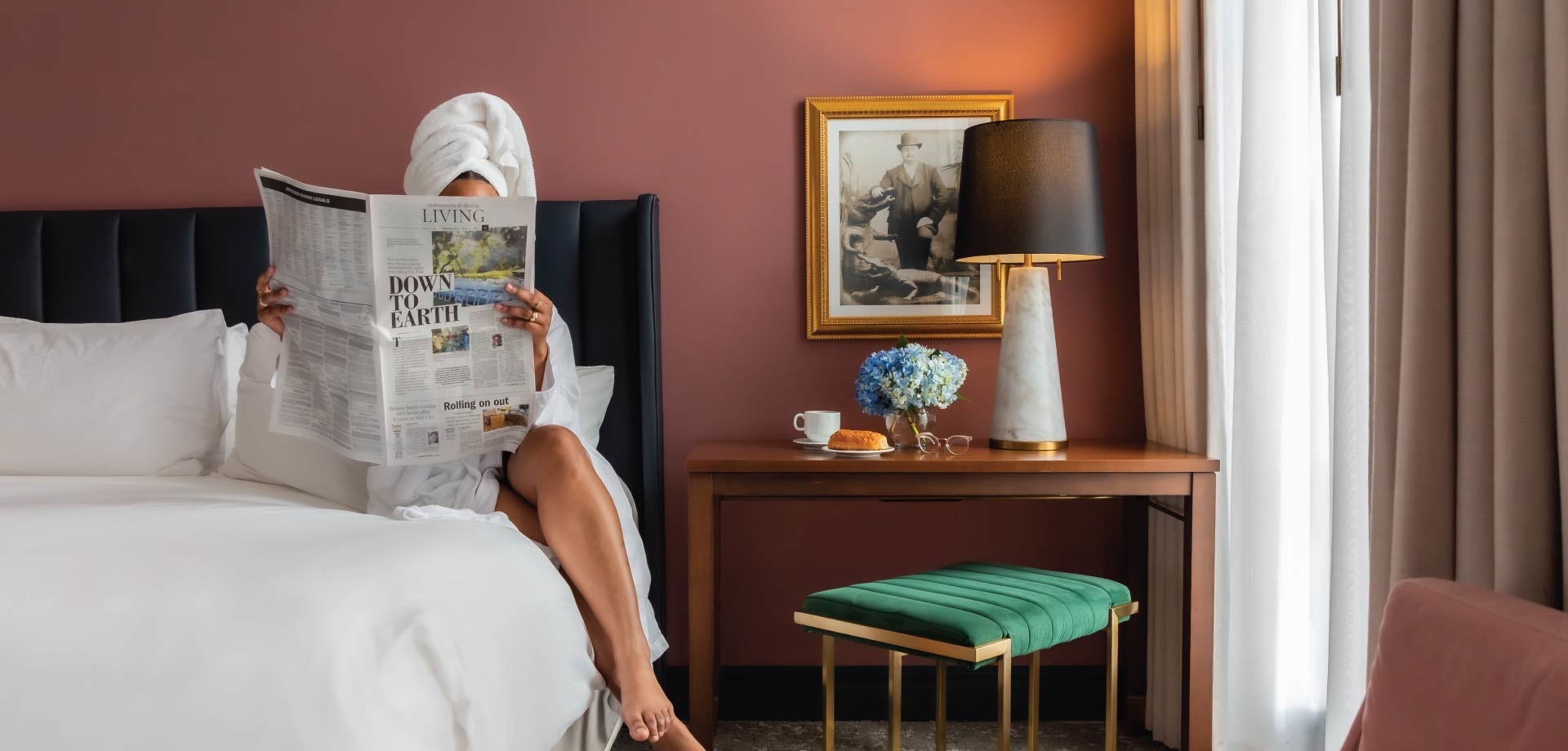 woman sitting on hotel bed with robe and towel on her head looking at a magazine with pink walls and green and blue accents