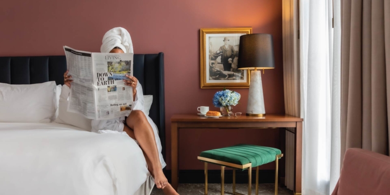 woman sitting on hotel bed with robe and towel on her head looking at a magazine with pink walls and green and blue accents