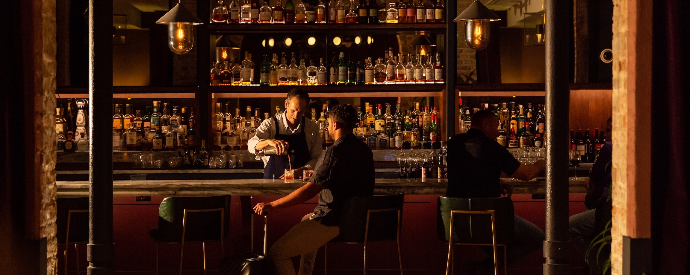 Man sitting at the bar near his suitcase with a bartender pouring a cocktail
